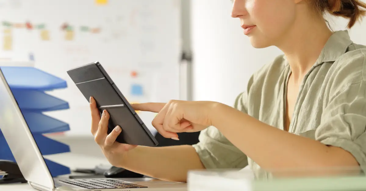 Woman sitting at desk with laptop, using tablet in front of screen